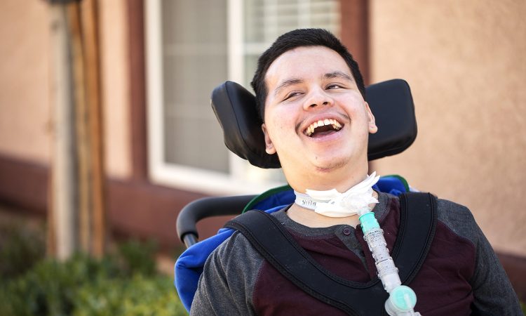 A young man with a tracheostomy tube is sitting outside his house in his wheelchair and smiling.