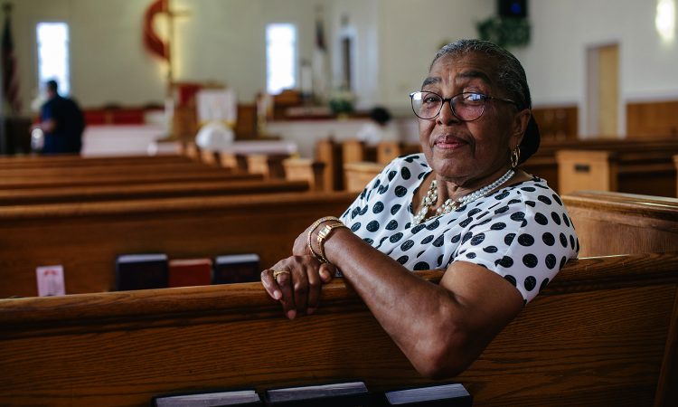 An elder African-American woman is sitting in a church pew and looking at the camera.