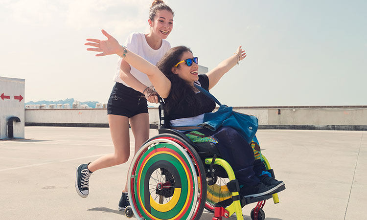 Female caucasian caregiver pushing female caucasian in a wheelchair with rainbow wheels. They are laughing and smiling.