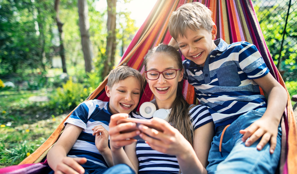 young adult girl with her two brothers on either side of her sitting on a hammock and smiling at her phone