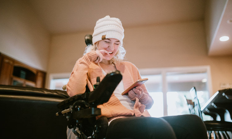 young woman in winter hat sitting in wheelchair and smiling at something on her phone