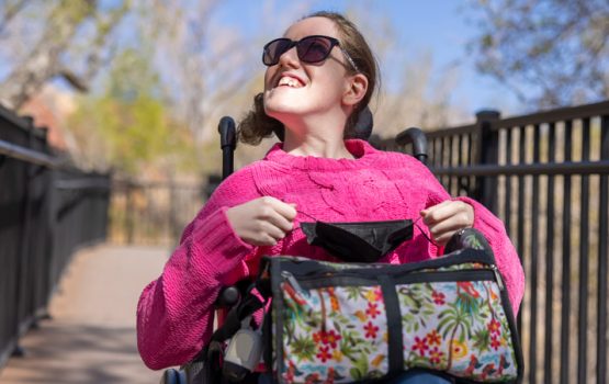 young woman with disabilities in wheelchair with hot pink sweater on and carrying a bag