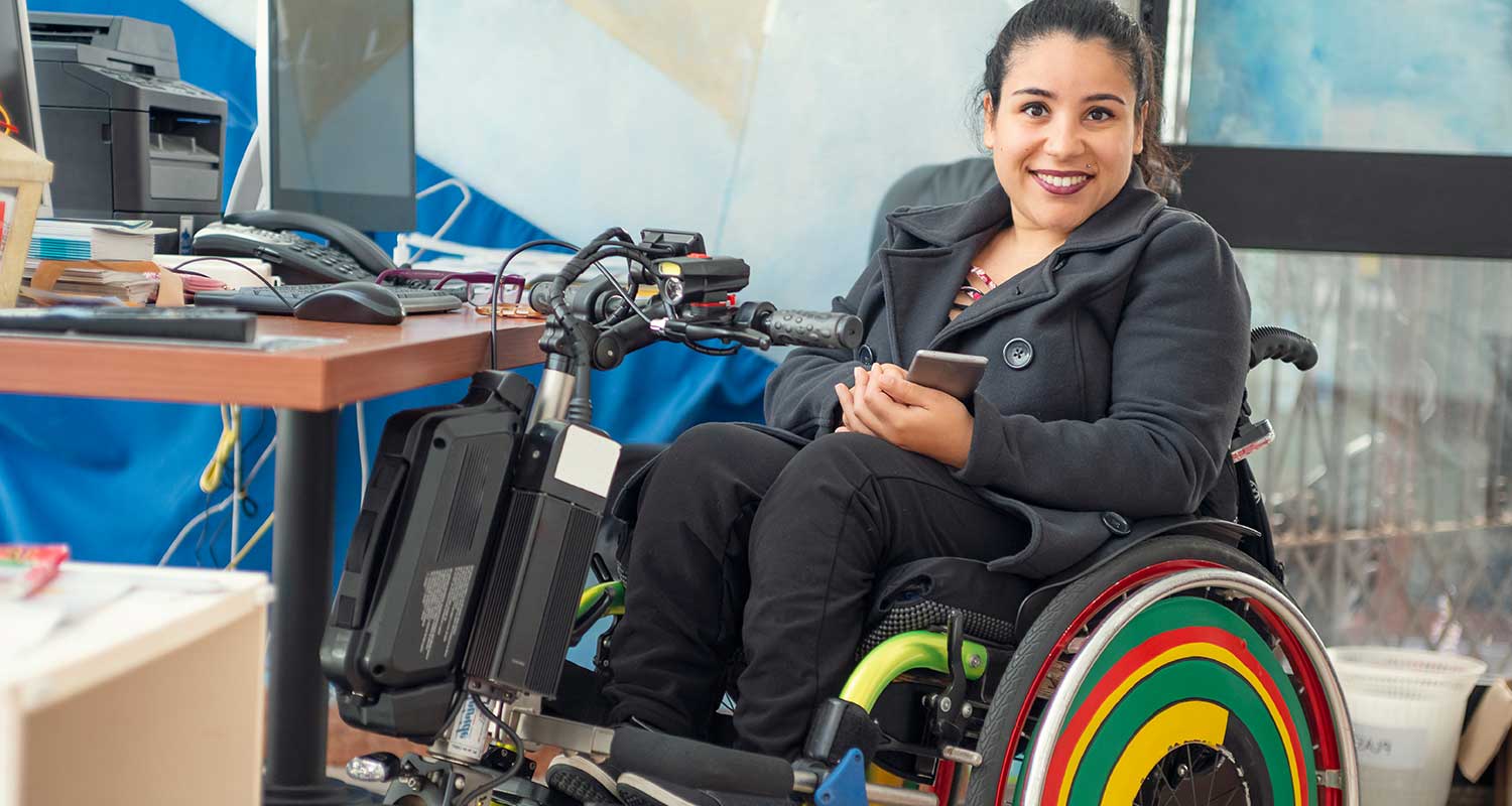 Woman in wheelchair with rainbow wheels at work desk, smiling at camera