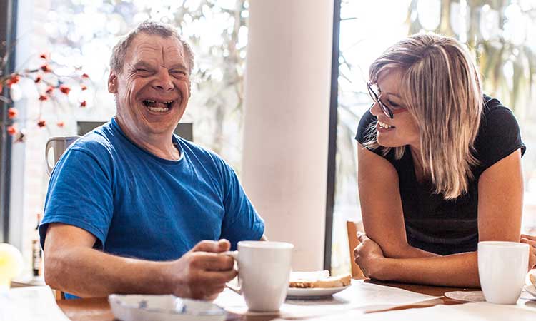 A woman is having coffee and laughing with a man with disabilities in their home.