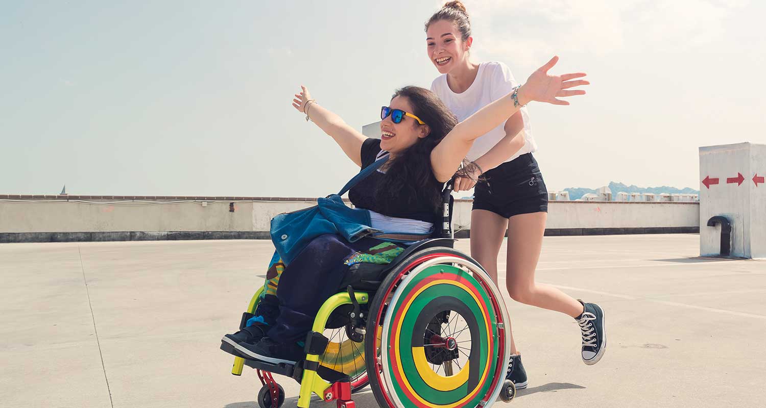 A female Caucasian caregiver is pushing a Caucasian woman in a wheelchair. The woman in the wheelchair has her arms raised. They are laughing and having fun.