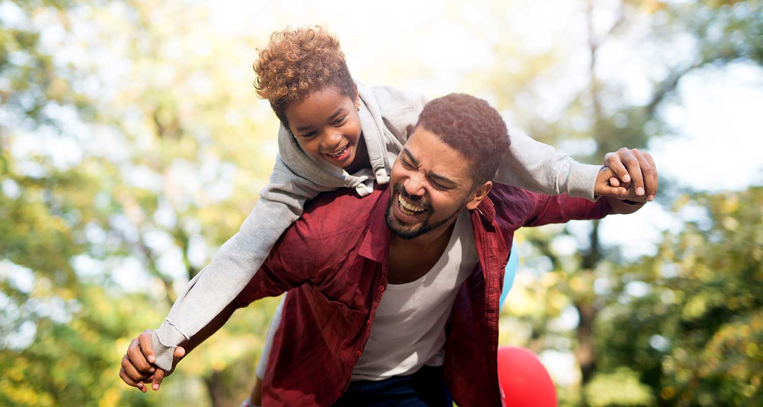 African-American father carrying his daughter on his back. They are in the park smiling and having fun.