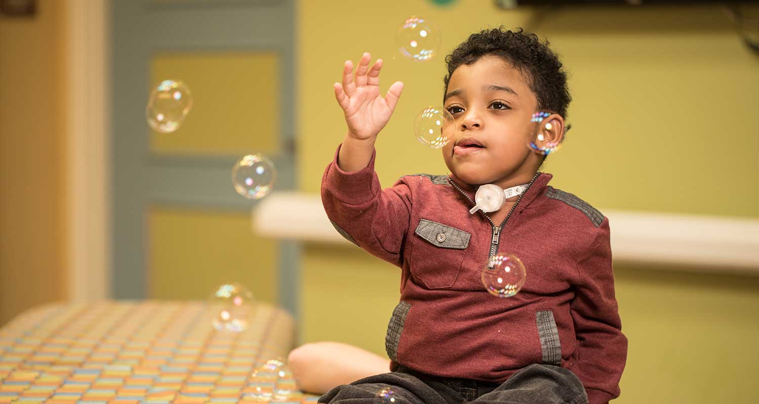 Toddler with tracheostomy tube sitting on couch and playing with bubbles