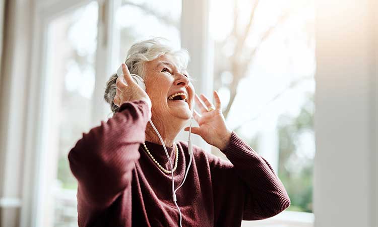 Senior Caucasian woman listening to music on headphones and smiling in front of a sunny window