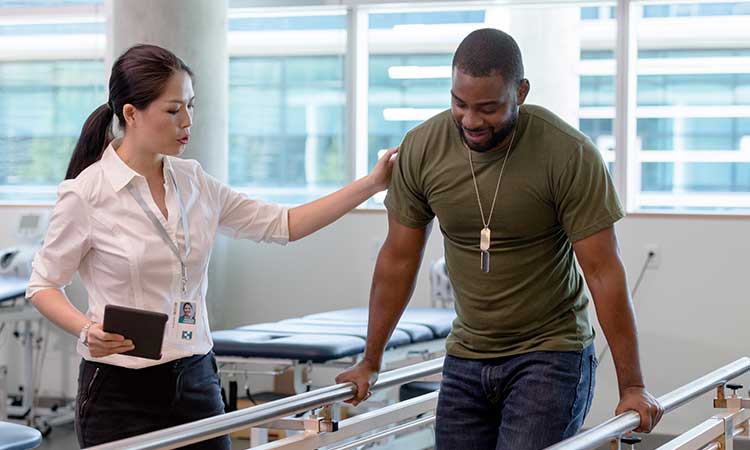 Female physical therapist is helping male veteran patient use parallel bars during rehabilitation