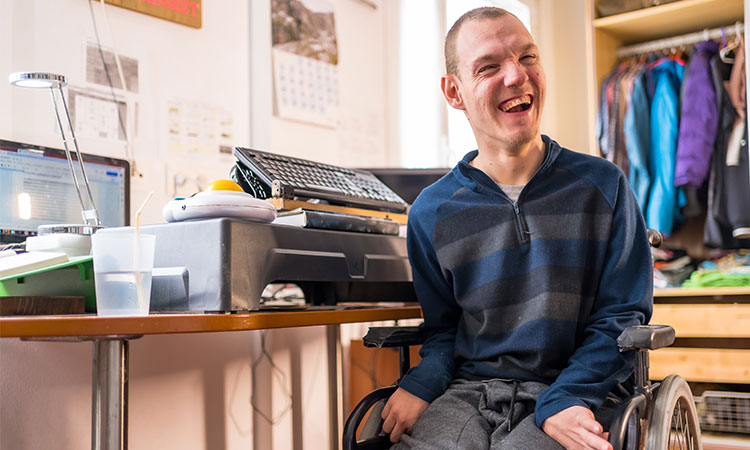 Young man sitting in a wheelchair at a desk in his bedroom and smiling