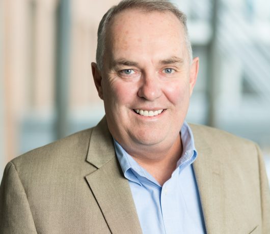 Bill Duffy is a man wearing a tan jacket and blue shirt and smiling at the camera.