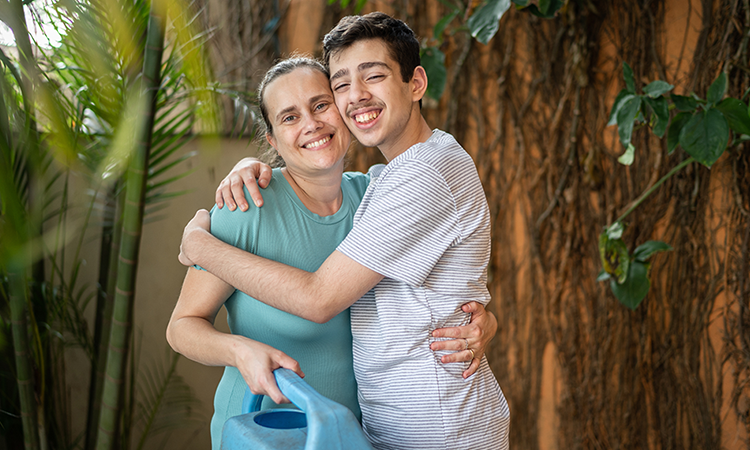 Mother with a watering can hugging her young adult son outside in their garden