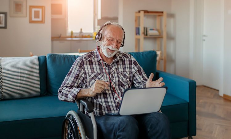 Senior adult man in a wheelchair listening to music on his laptop with headphones on and smiling.